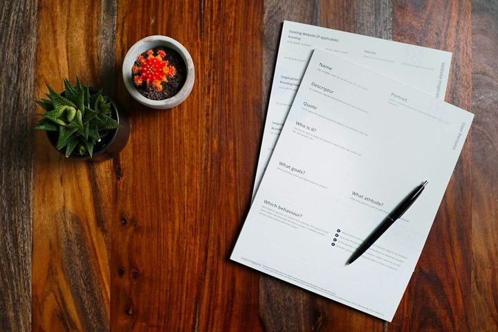 A multi-page printed form fanned across a dark wooden table, the top sheet showing ruled section headings and mostly blank fields, a pen lying across it, two small potted plants at the edge.
