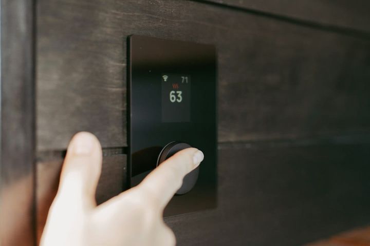 A hand reaching up to a wall-mounted digital thermostat set into dark wood panelling. The small display reads 63 with a heating indicator lit above it.