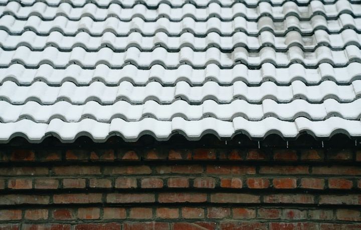 A tiled roof under an even layer of snow above a brick wall in flat overcast light, the tiles visible only as ridges through the covering and the roof surface itself hidden.