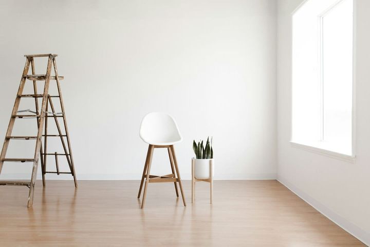 A nearly empty room with bare white walls and a pale wood floor, lit by one window. A wooden stepladder stands by a single stool and a potted plant. No people are in frame.