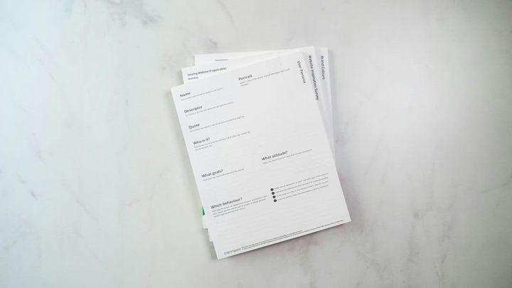 A small stack of printed report pages fanned on a pale stone worktop in flat daylight, the top sheet showing ruled section headings and short field labels with nothing filled in.