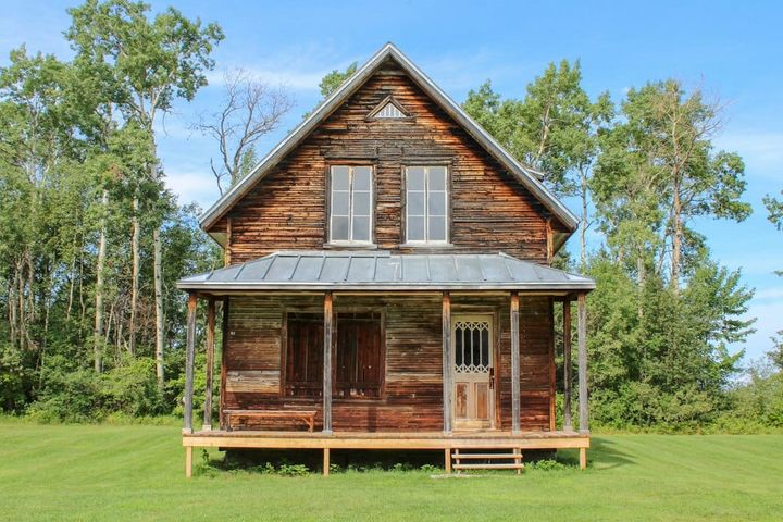 A weathered wooden cottage with a steep front gable, two upper windows and a full-width covered porch, standing on a mown lawn against trees.