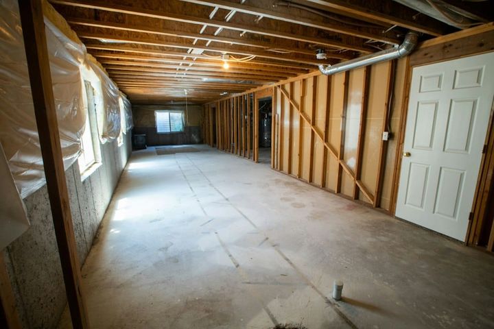 An unfinished basement with exposed floor joists, open stud framing along one wall, a metal duct overhead and a bare concrete floor.