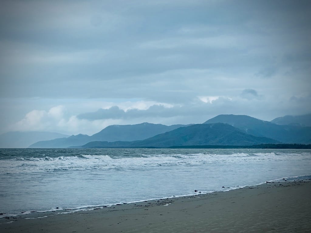 A beach on a gray day, with low breakers in the front and gray-green hills in the distance