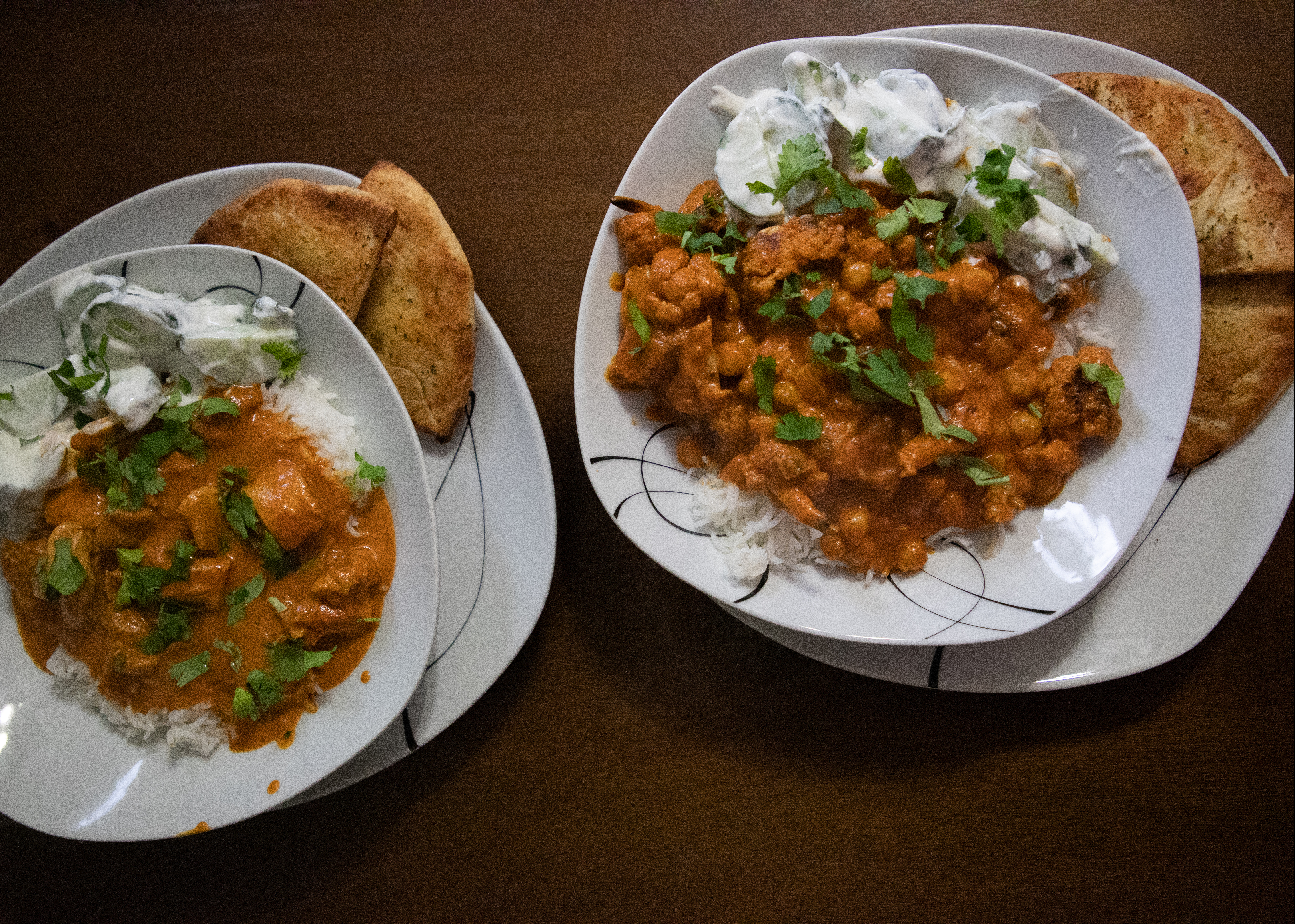 Two dishes of curry over a bed of white rice with garlic naan and cucumber raita on the side