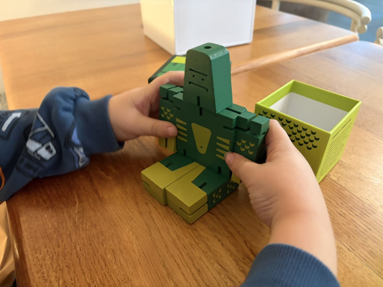 A child's hands holding a green and yellow wooden Cubebot robot at a café table, with its box beside it.