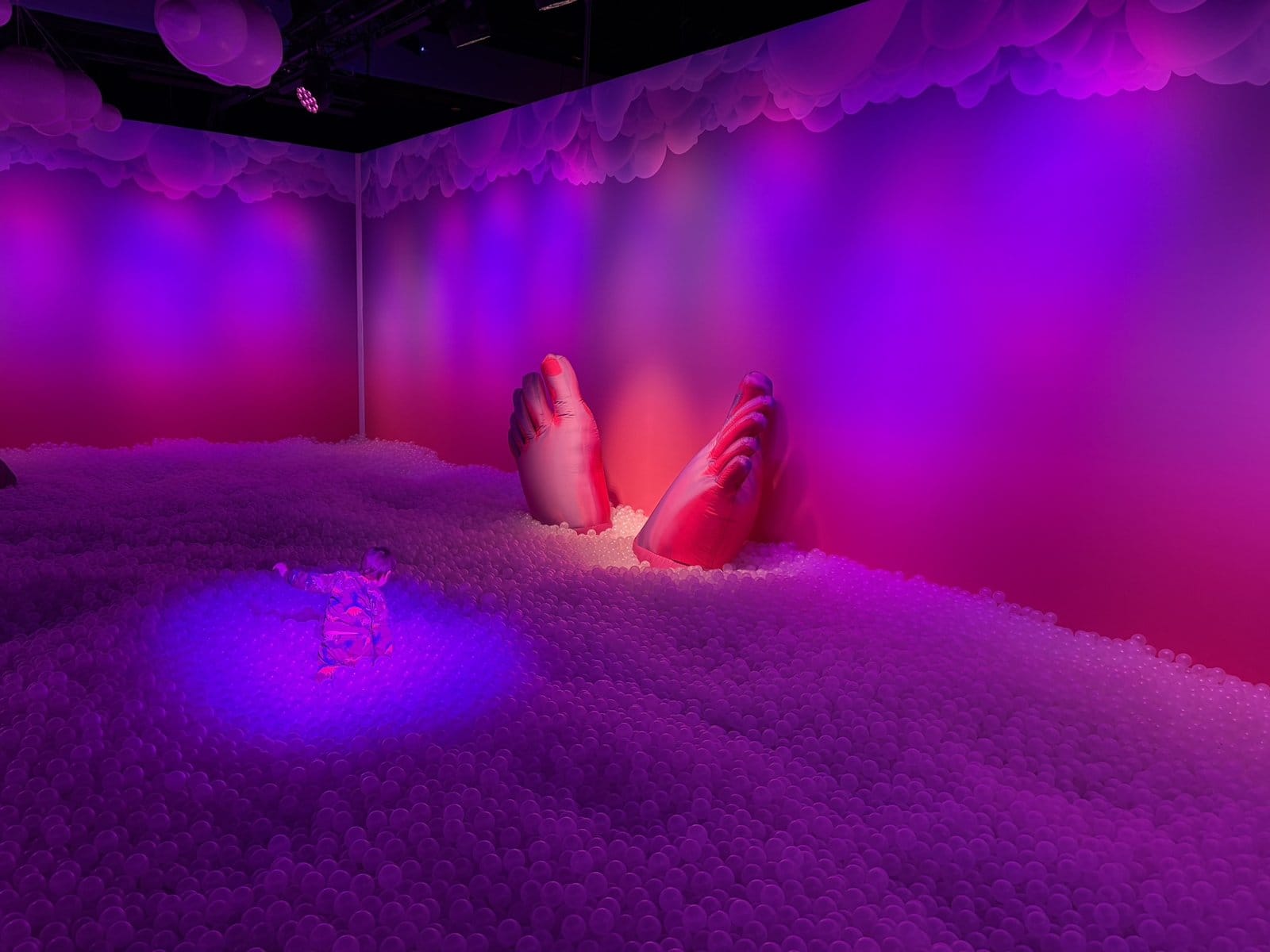 Giant feet emerging from a deep pit of pale pink balls in a purple-lit room. A young boy, viewed from behind, is wading through the balls.