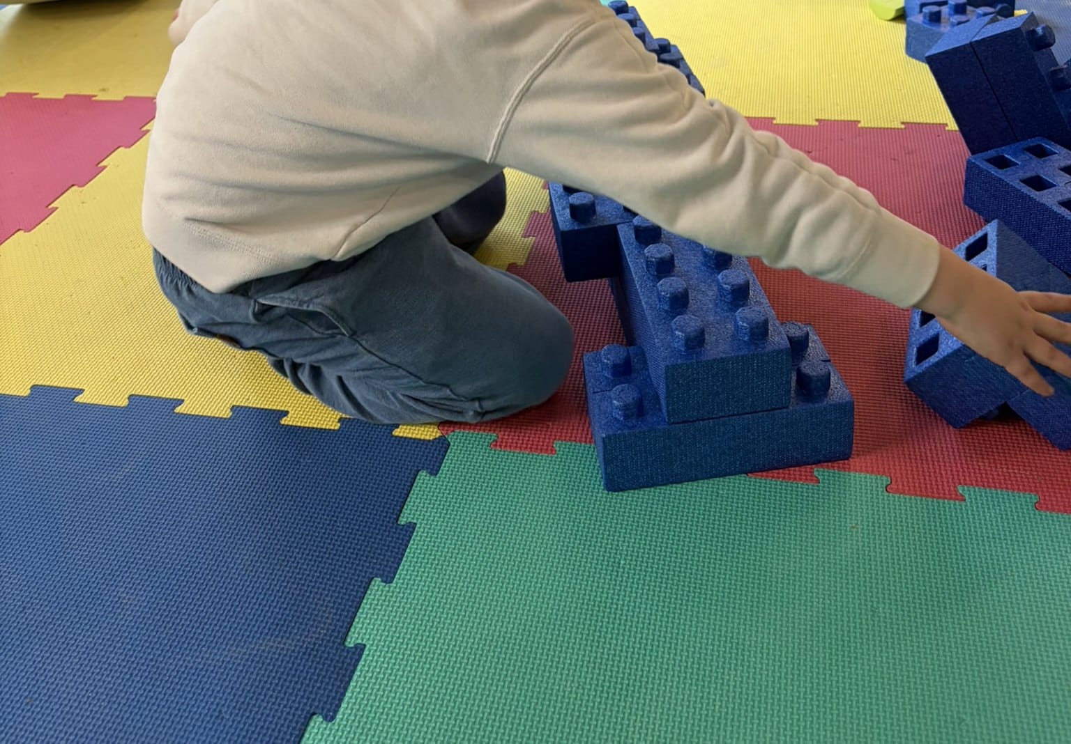 A child kneeling on a foam mat floor building with oversized blue plastic blocks.