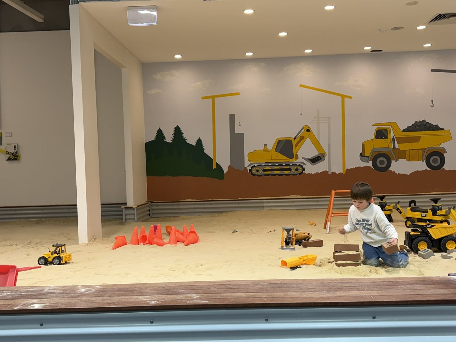 A child playing alone in a wide indoor sandpit in front of a painted mural of excavators and dump trucks.