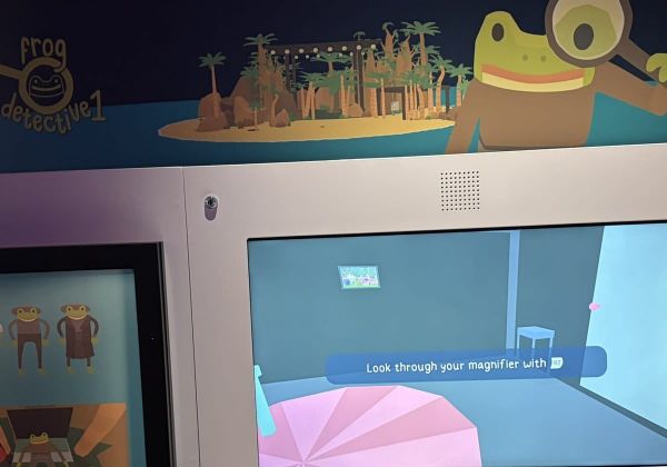 A videogame on a large screen in a museum games area, with illustrated frog artwork on the wall above.