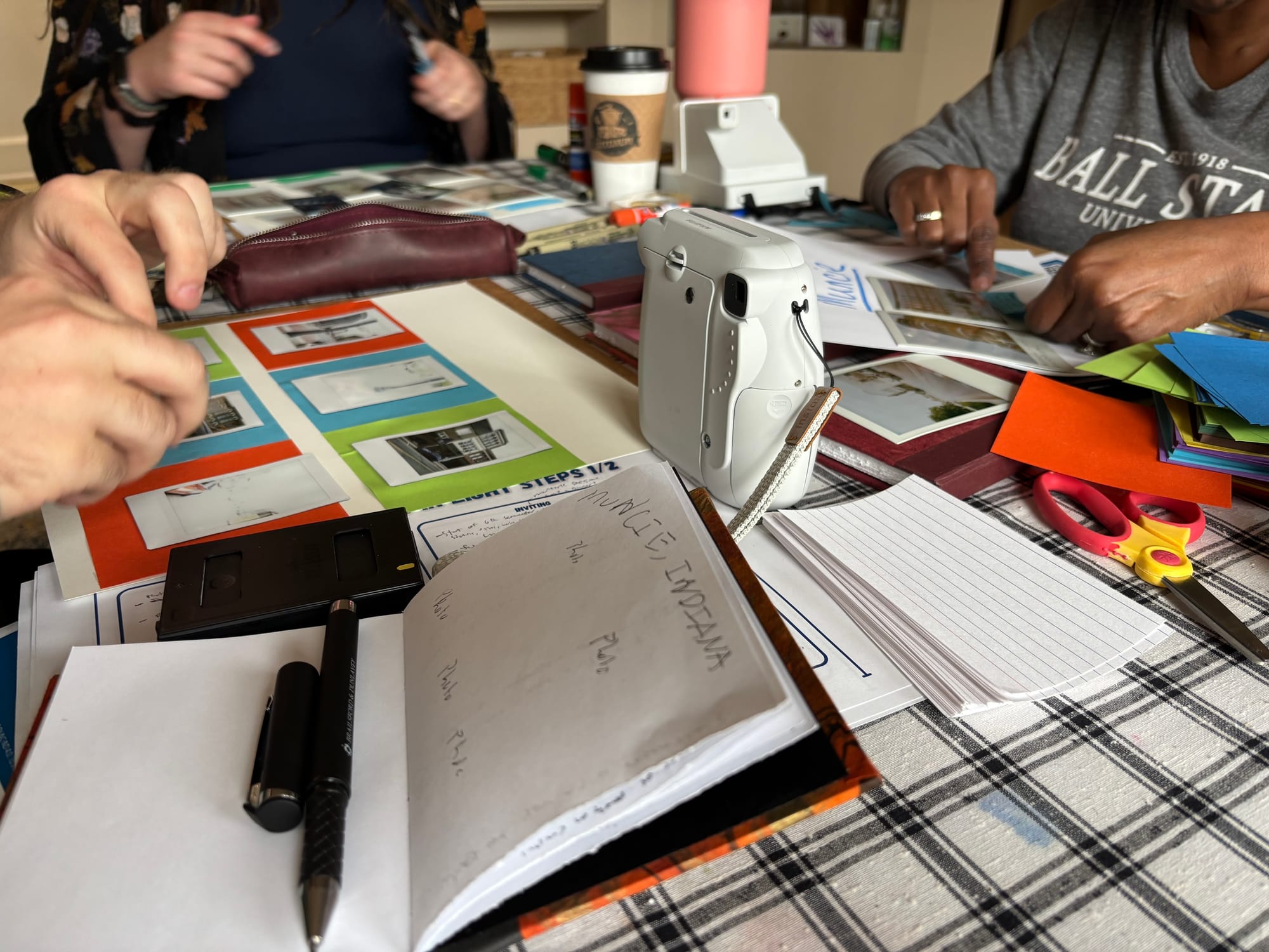 table full of scrapbooking supplies with three people actively working on scrapbook pages