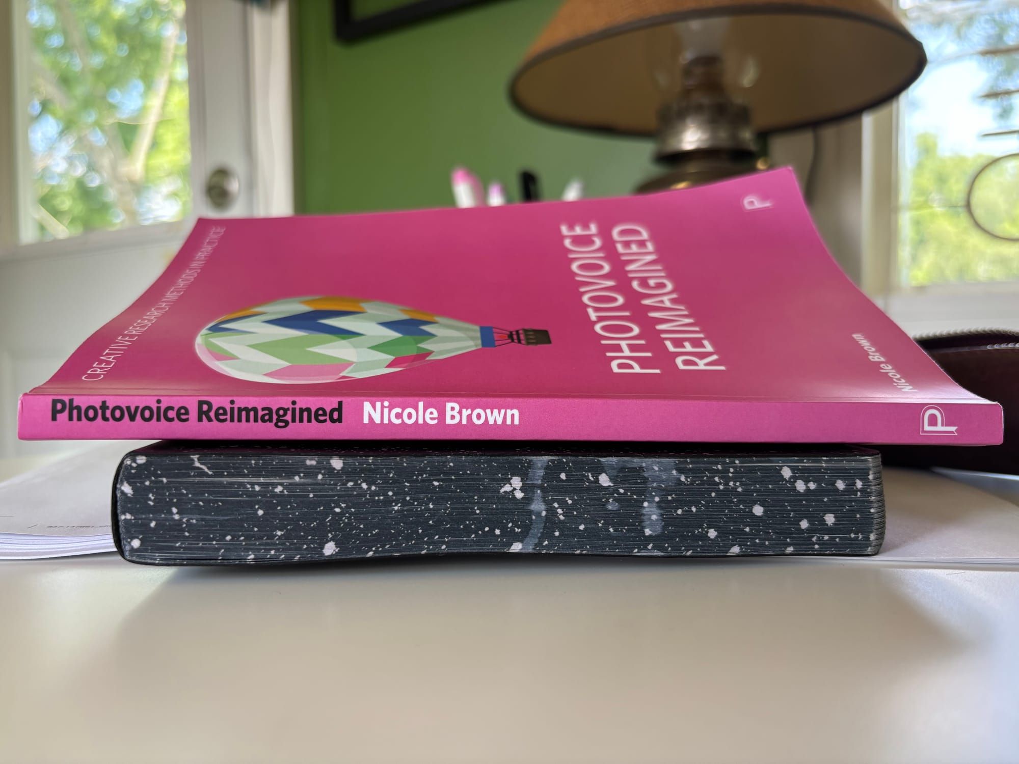a pink book with a hot air balloon on the cover titled Photovoice Reimagined by Nicole Brown sitting atop a journal on a white desk