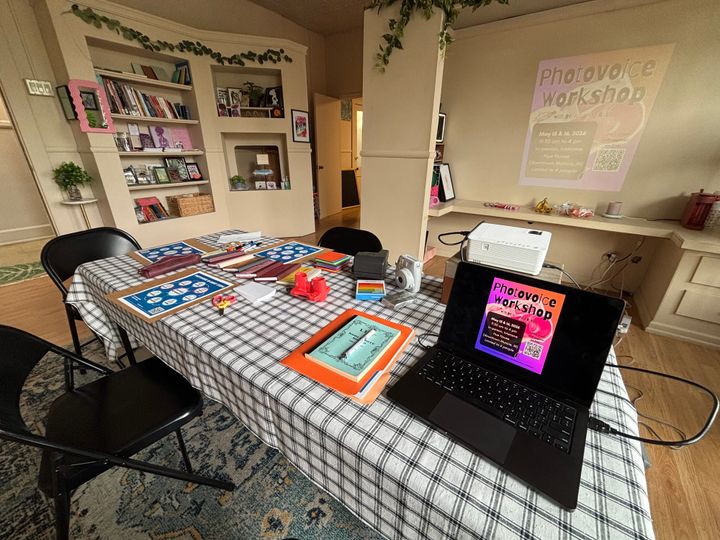 table in the middle of a room; atop the table are papers, Polaroid cameras, papers, notebooks, office supplies, and a laptop and projector.