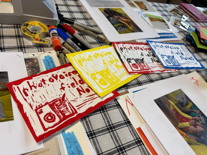 tabletop filled with markers, handouts, polaroid photographs, papers, a roll of tape, a glue stick, and four linocut prints containing the text "photovoice field notes"