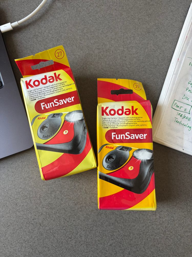 two disposable cameras on a desk with a laptop and notebook on either side