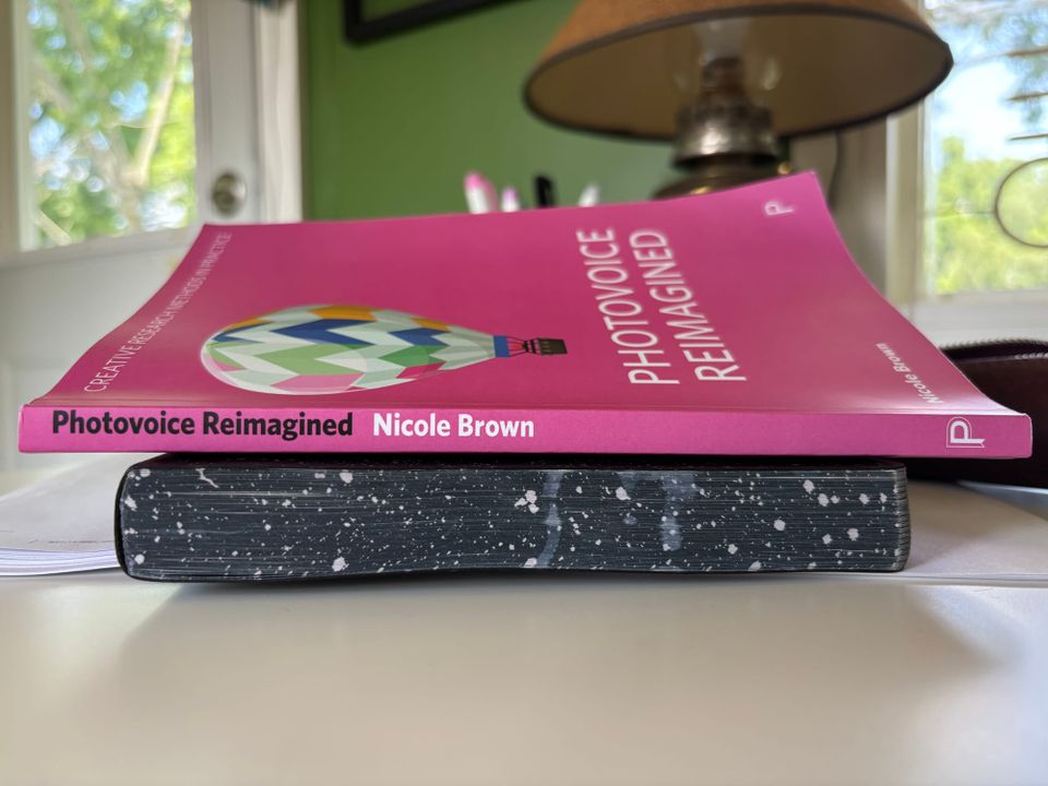 a pink book with a hot air balloon on the cover titled Photovoice Reimagined by Nicole Brown sitting atop a journal on a white desk