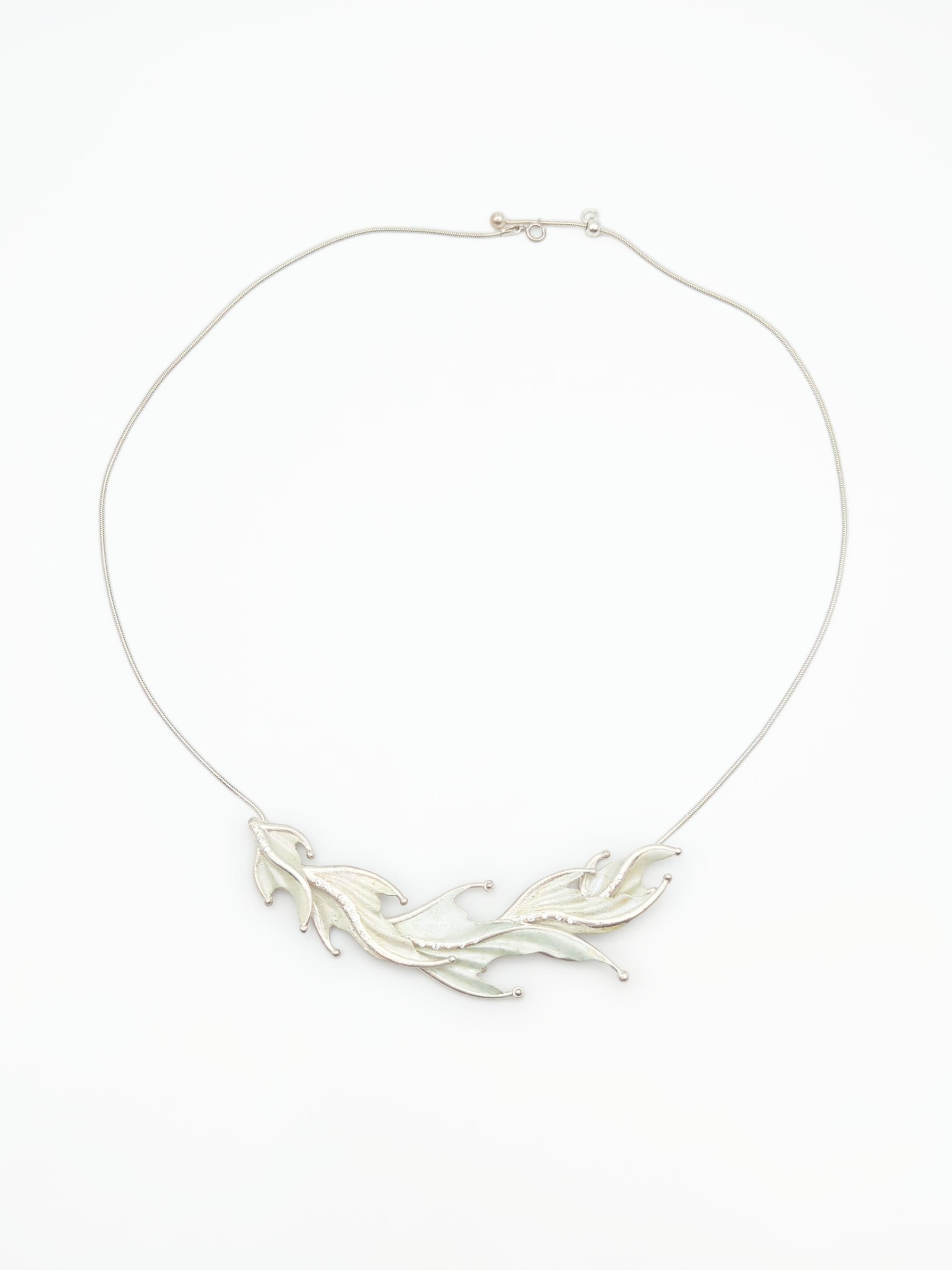 The five 'fins' of the necklace are graduated in size, with the two smallest on the outsides and the largest in the middle. In this photo they flow left-to-right, each one slightly overlaps the next. Behind either side a round smooth chain is attached. The chain has an adjustable clasp at the end.