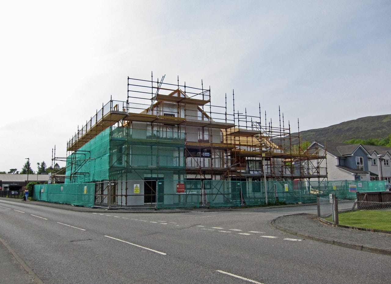 A block of new council homes in green safety netting and scaffolding beside a road in Portree, hills behind