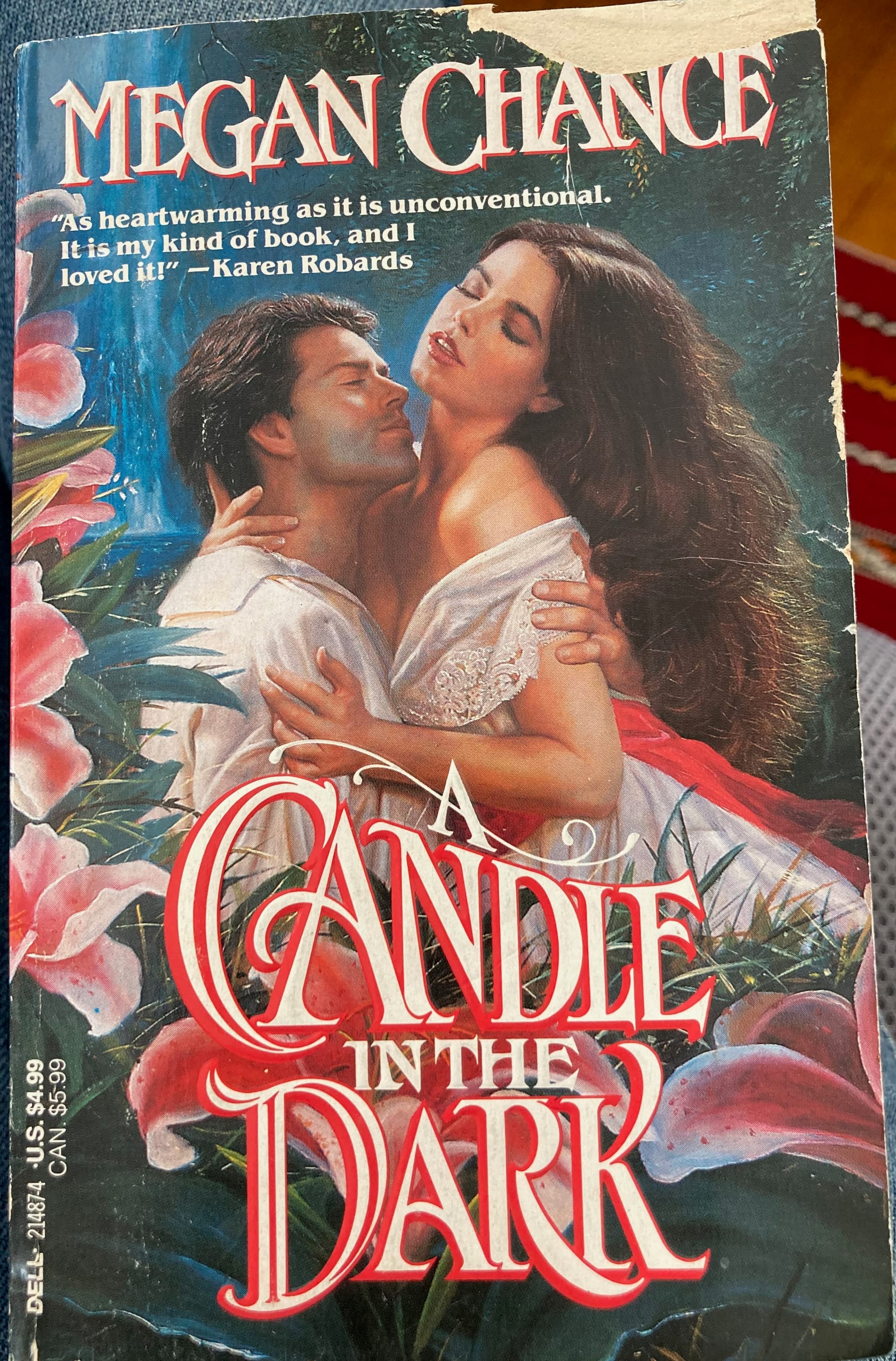 A slightly worn paperback copy of Megan Chance's A Candle in the Dark. The cover illustration shows a white man pulling a white woman into his lap to embrace her. Her white lace dress is falling off her shoulder. There is a border of painted lilies. They're pink and sexually suggestive. The title text is white with a red border and big swooping capitals.