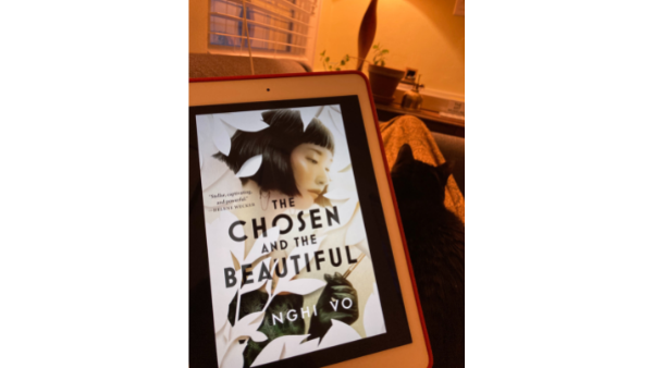 An ereader showing the cover of Nghi Vo's The Chosen and the Beautiful, which features an Asian woman with short hair dressed in 1920s clothes among white flowers. In the background of the photo is a black cat.