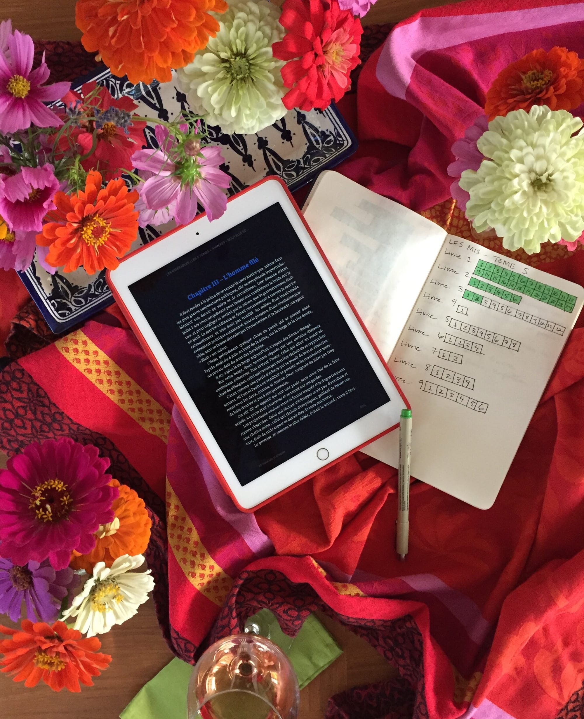 A photo of some zinnias, cosmos, and lavender from my yard in flower arrangements, on top of a colorful tablecloth with a glass of wine, a notebook, a pen, and my iPad with the screen showing a page from Les Mis.&nbsp;Also, I guess I do #bookstagram…