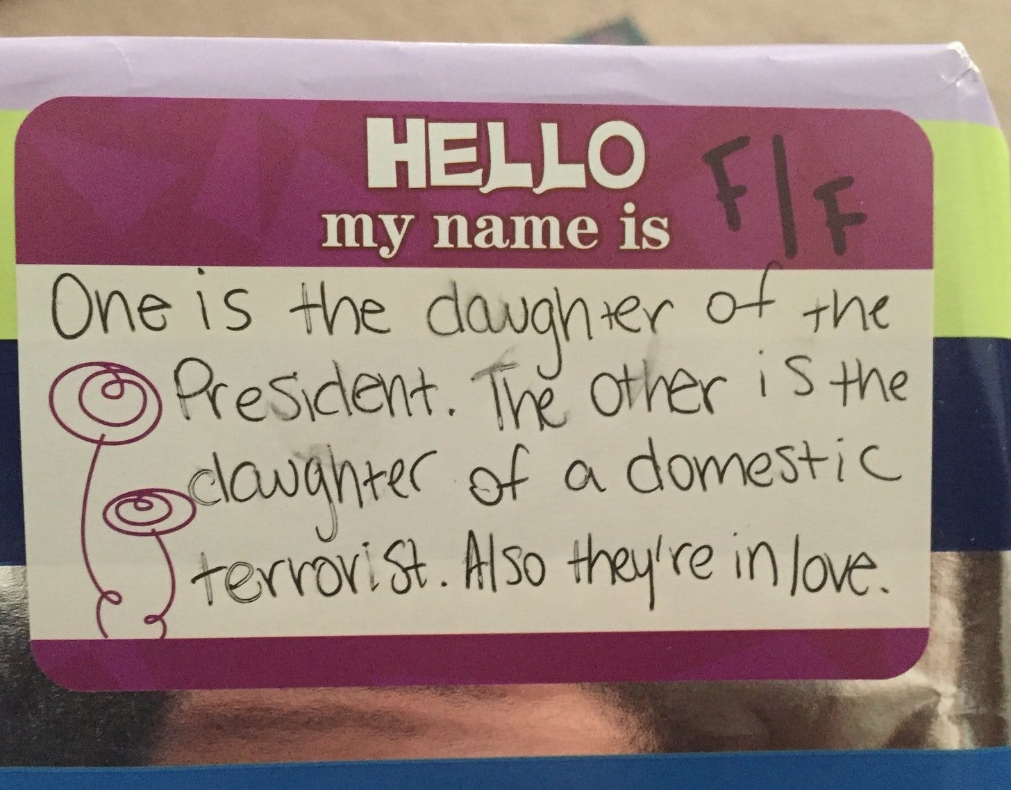 The image is of a book in shiny silver wrapping paper, and the handwritten nametag on the paper says “Hello, my name is… F/F. One is the daughter of the President. The other is the daughter of a domestic terrorist. Also they’re in love.”