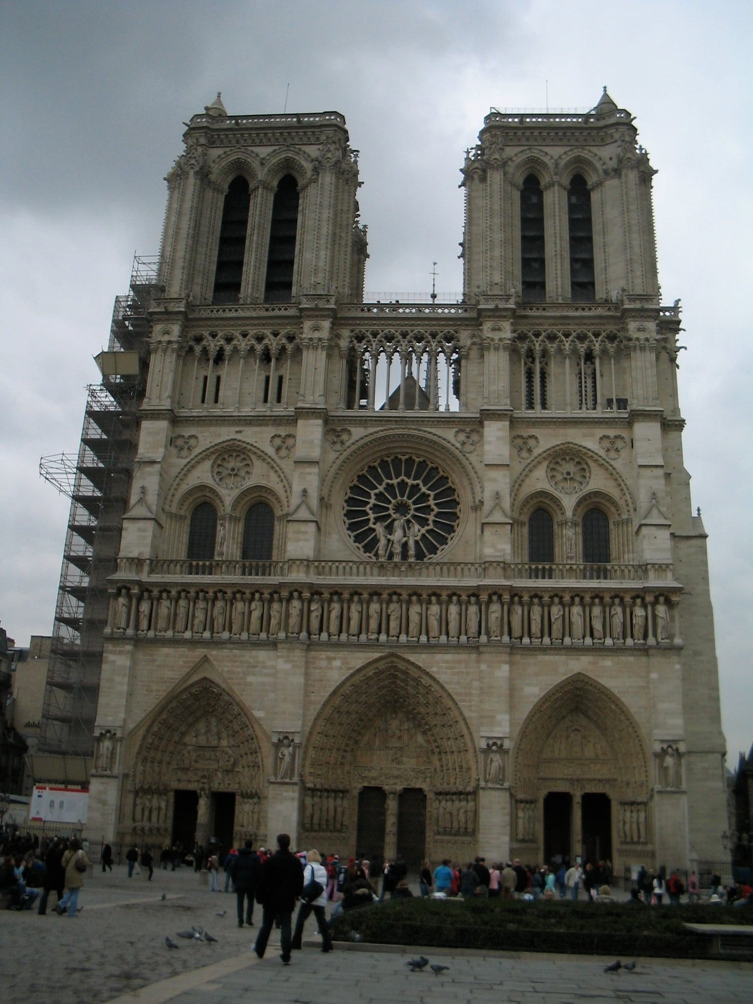 The facade of Notre Dame de Paris on a cloudy day. The cathedral is framed slightly crooked in the photo and there are tourists milling around in front of it.