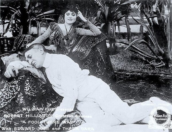 a black and white photo of a woman lounging smugly on a chaise longue while a man lies limp and supplicant with his head in her lap