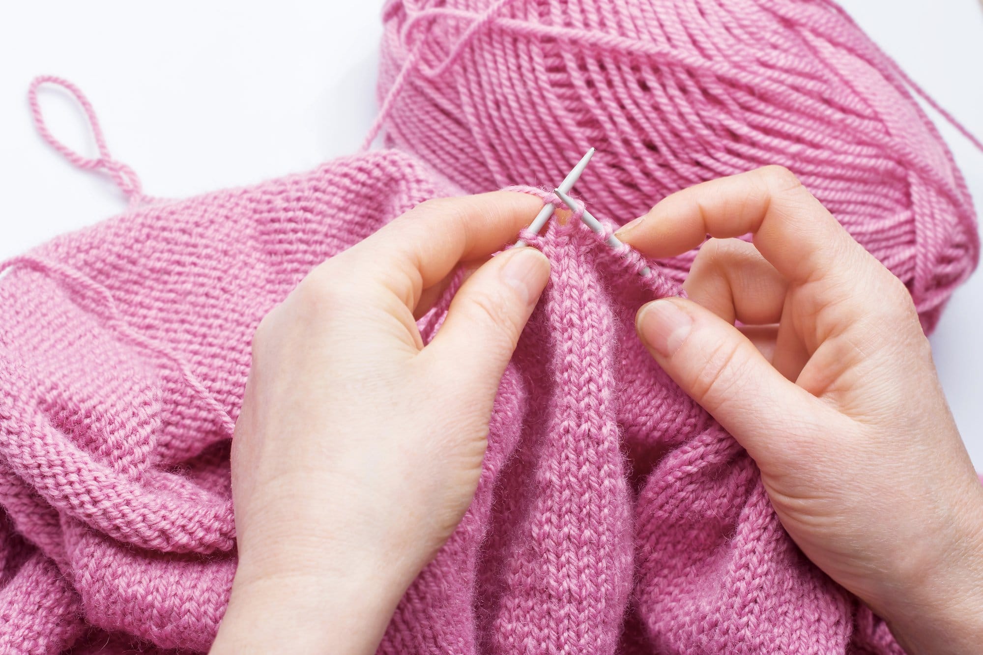 Two white hands holding a pair of knitting needles with many stitches of pink yarn on them. There is a long length of knitting coming off the needles and a skein of yarn in the background.