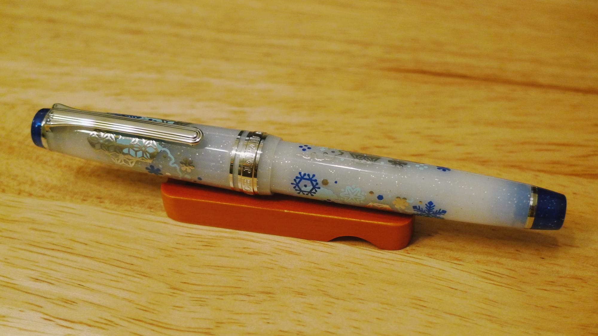 Opaque milky white fountain pen with silver trim, blue finials, and a glittery blue and silver snowflake design sitting on a small wooden pen rest