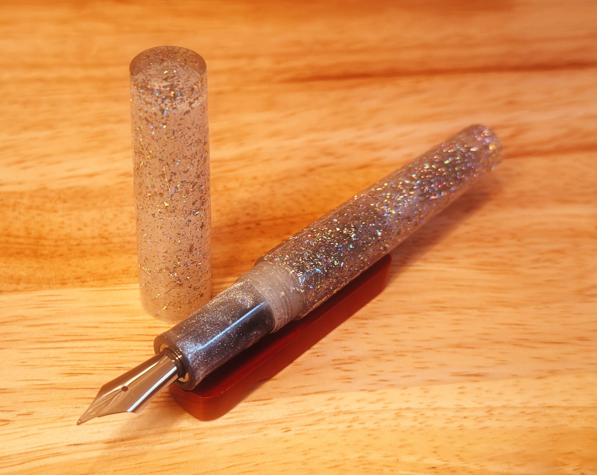 Uncapped fountain pen made of a translucent resin filled with chunky holographic glitter, and a grip section made of a silver resin filled with silver shimmer sitting on a wooden pen rest