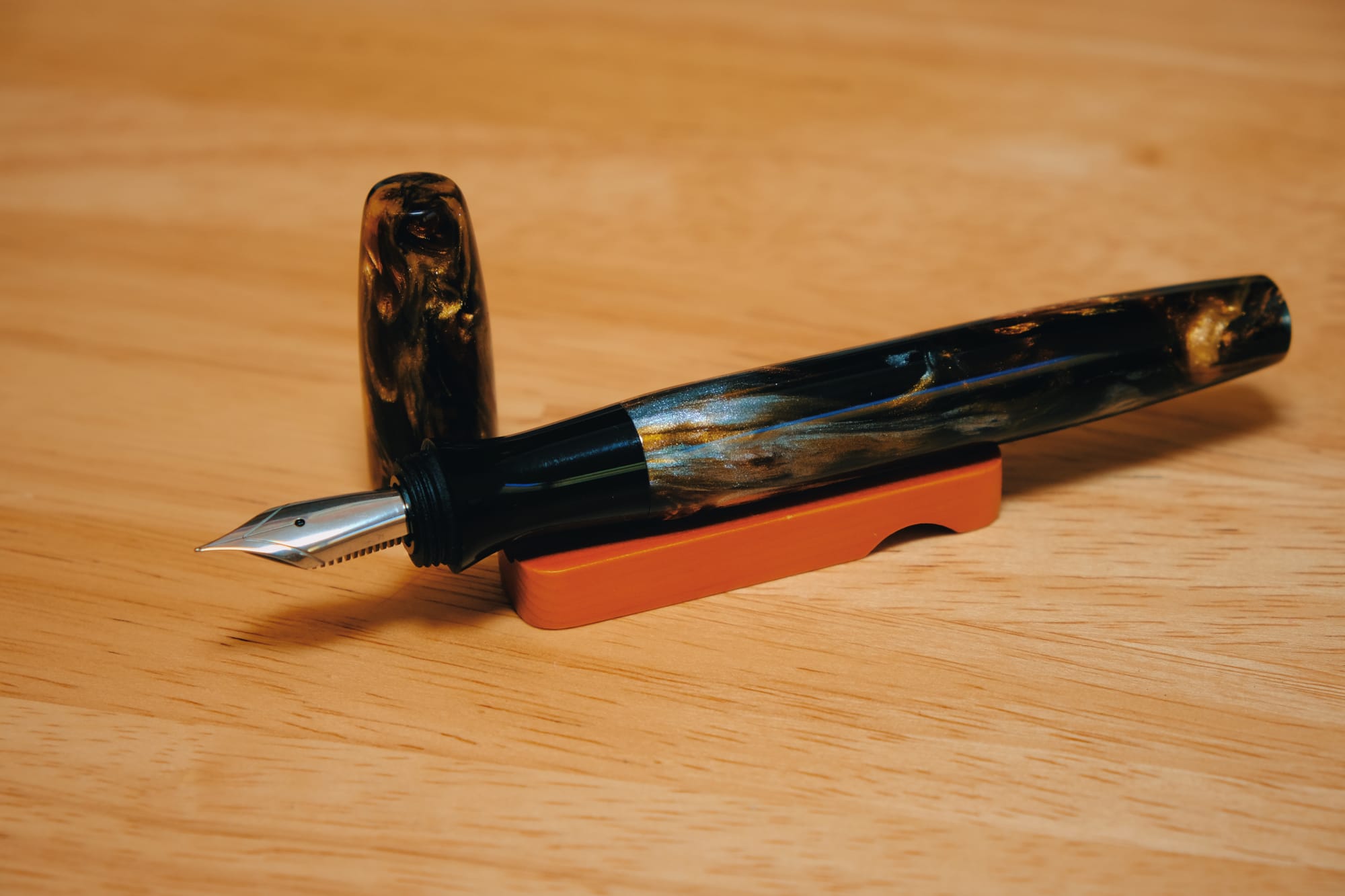 A fountain pen made out of a dark resin with swirls of golden brown and silver shimmer, sitting uncapped on a small wooden pen rest, it's cap sitting upright behind the pen grip