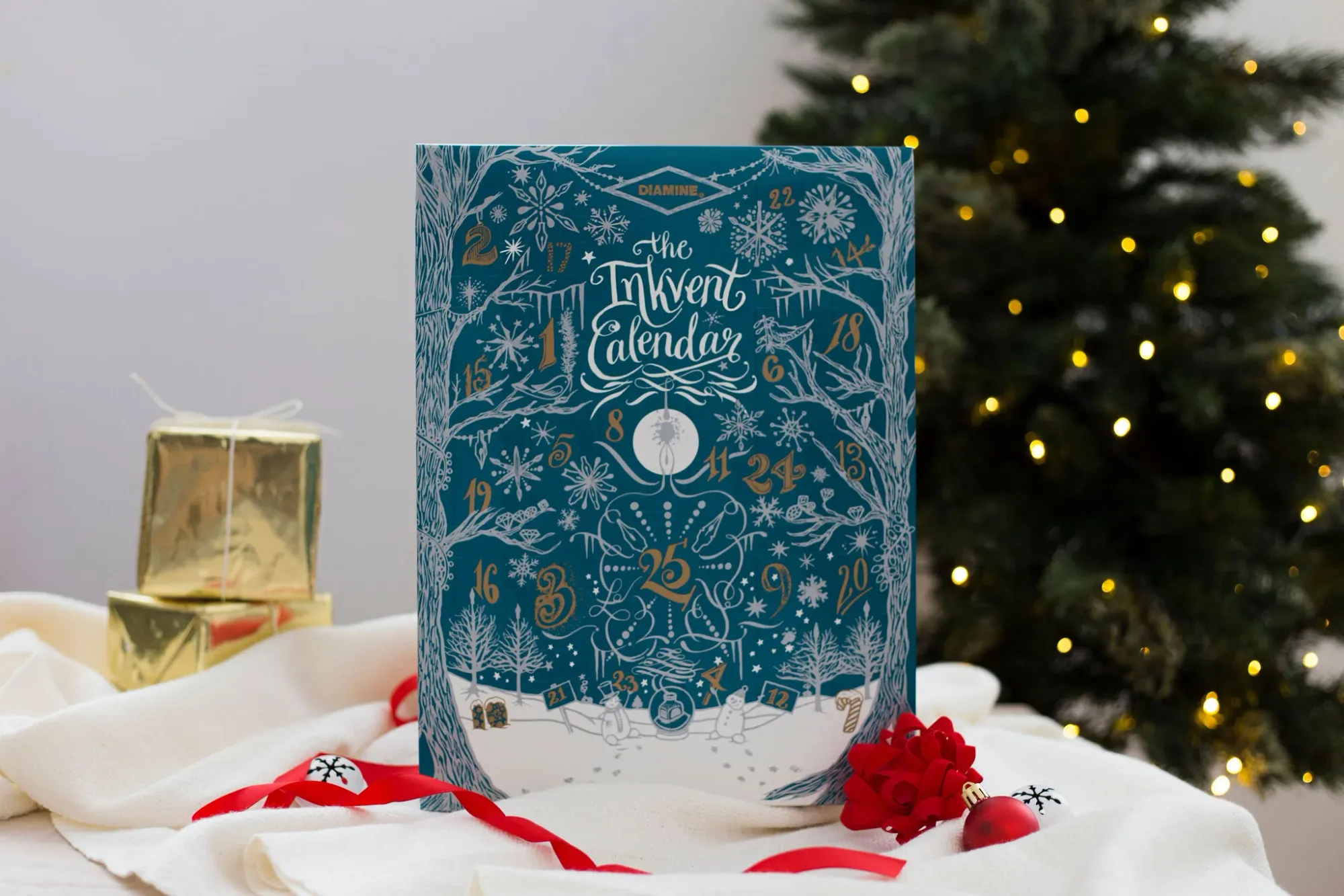 A teal box with holiday Advent tree illustrations, labeled "Diamine the Inkvent Calendar", with a lit Christmas tree and some gold-wrapped gifts in the background