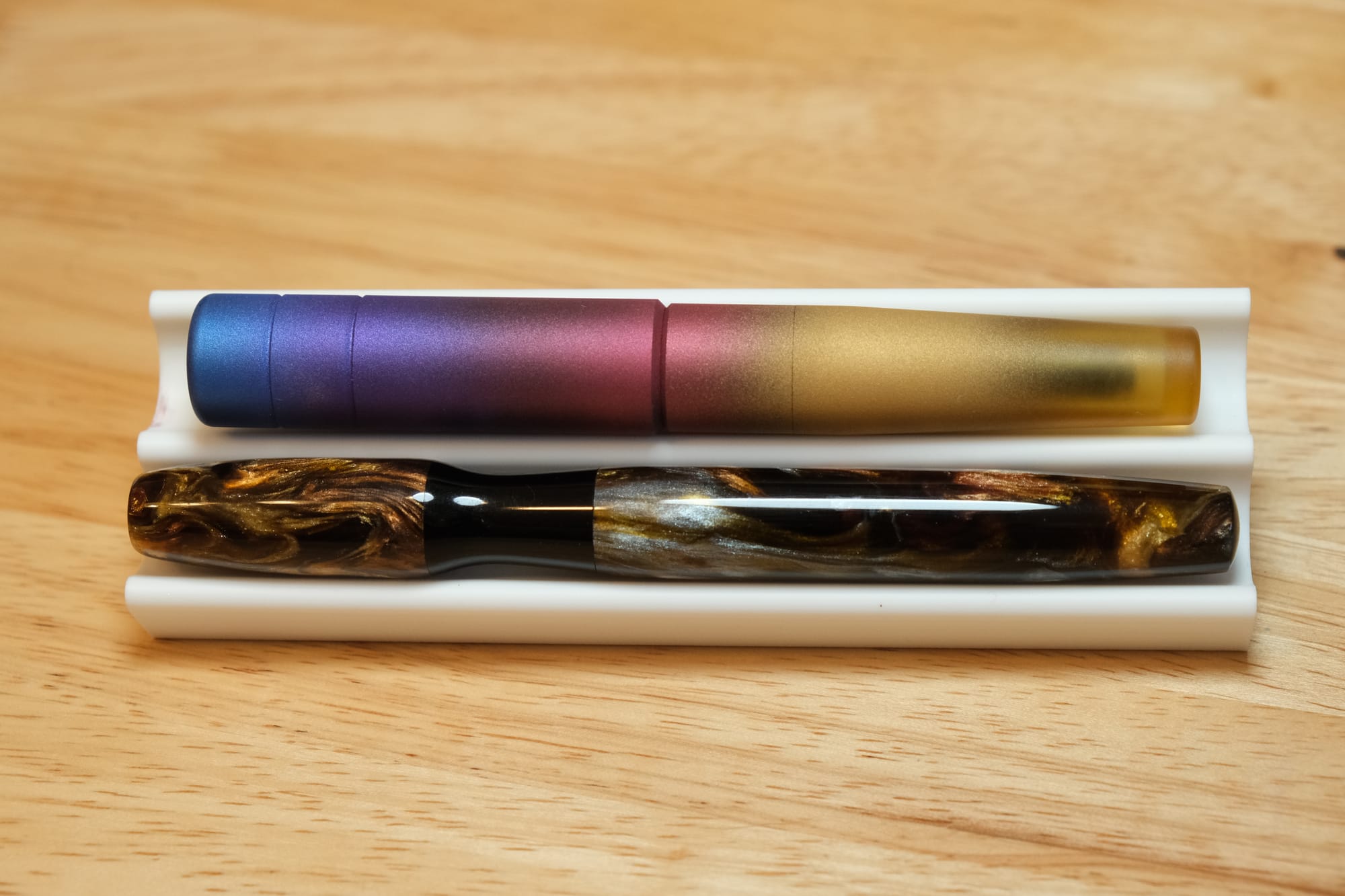 Two fountain pens sitting in a dual pen stand, vertically stacked; the top is an Ultem pen airbrushed in a sunset-colored gradient of blue, purple, dark pink, and yellow, the bottom pen is cigar-shaped with a black resin hourglass grip section, and a swirly black, gold, and silver shimmer resin for pen cap and body
