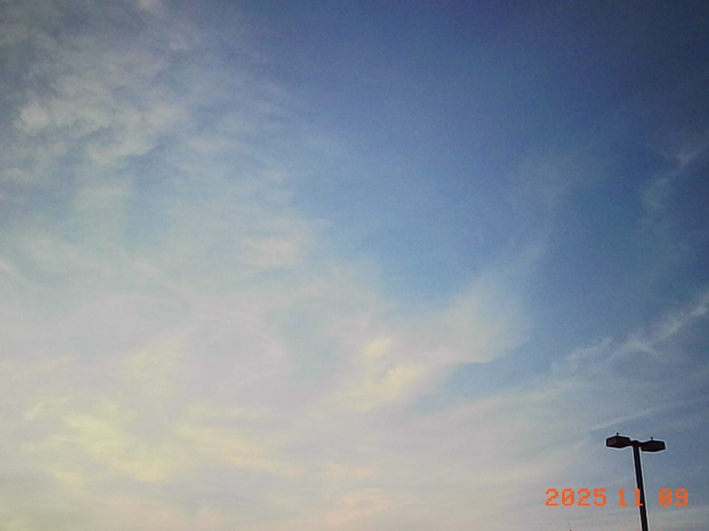 Low-res image of a partly cloudy sky close to sunset time, with the top of a parking lot light visible in lower right corner