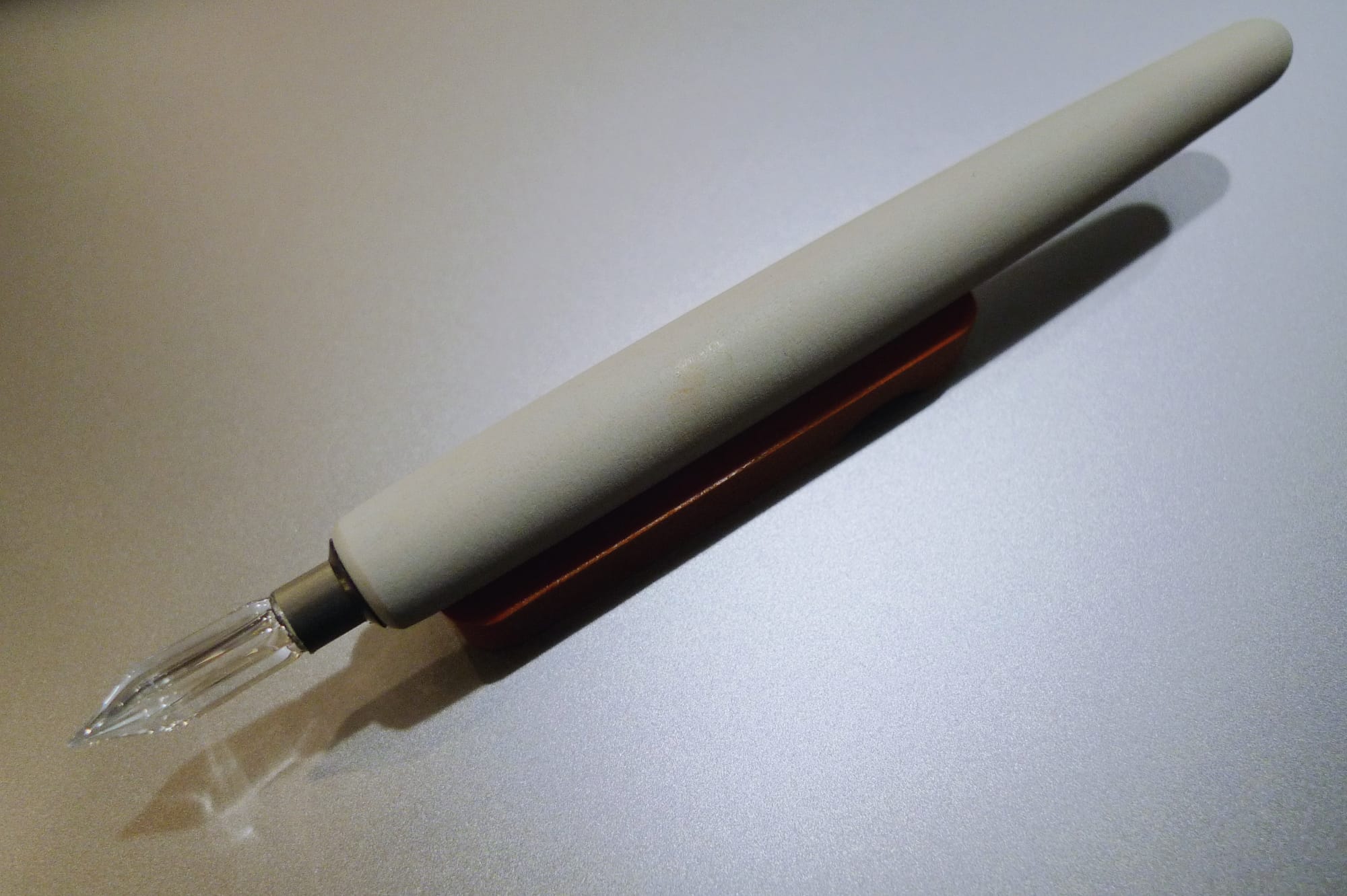 A glass nib fitted into an off-white/greige nib holder sitting on a small wooden pen rest on a silver surface