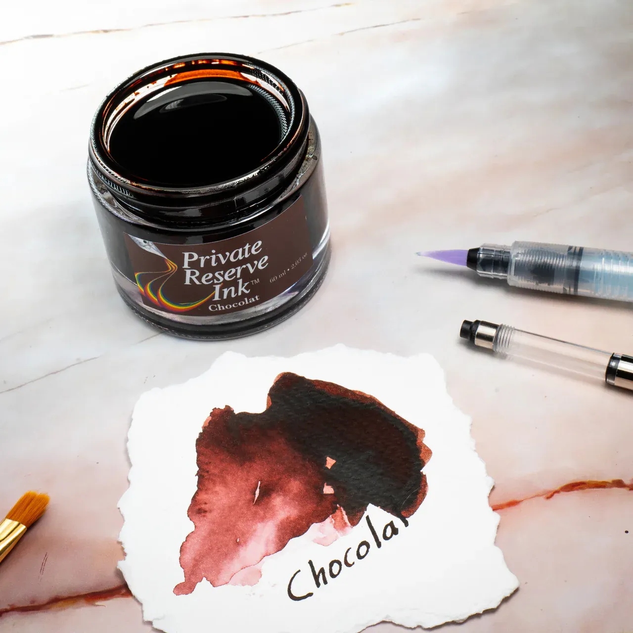 An open ink bottle with a wide opening and straight sides (basically a small jar), with a torn ink swatch sitting nearby on a table top with a water brush, cartridge converter, and a small paint brush on either side of the swatch paper