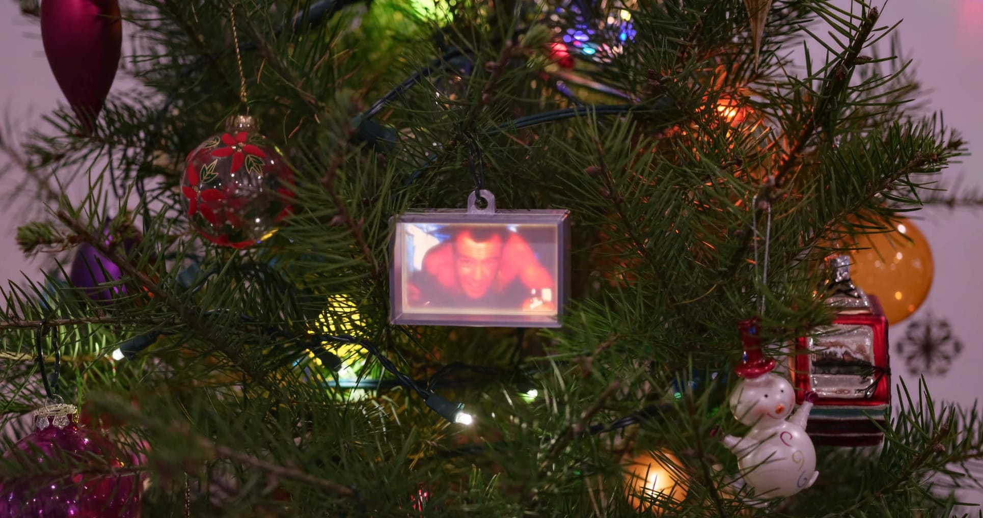 Close up of a Christmas tree with various regular ornaments surrounding a silver box ornament with a backlit, photograph-like scene of a man crawling in an air duct, holding a lighter