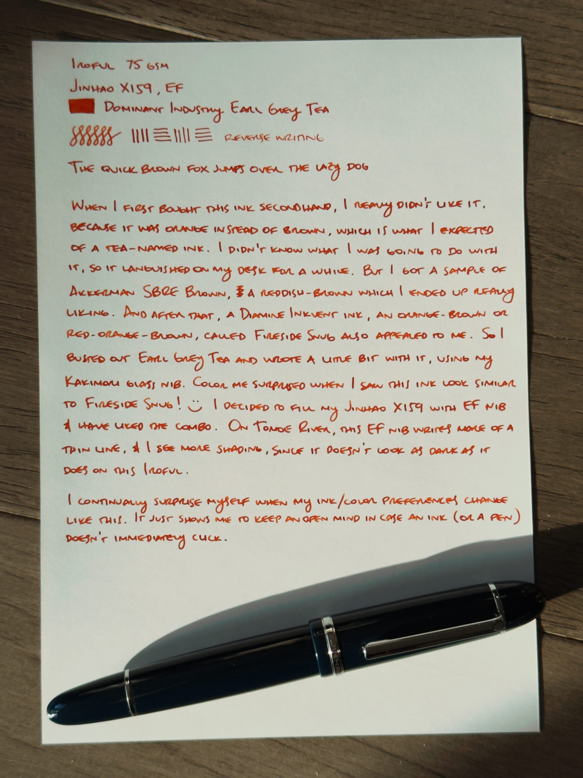 Writing sample using a red-orange ink on white paper, the dark blue, cigar-shaped fountain pen resting on the bottom of the sheet of paper