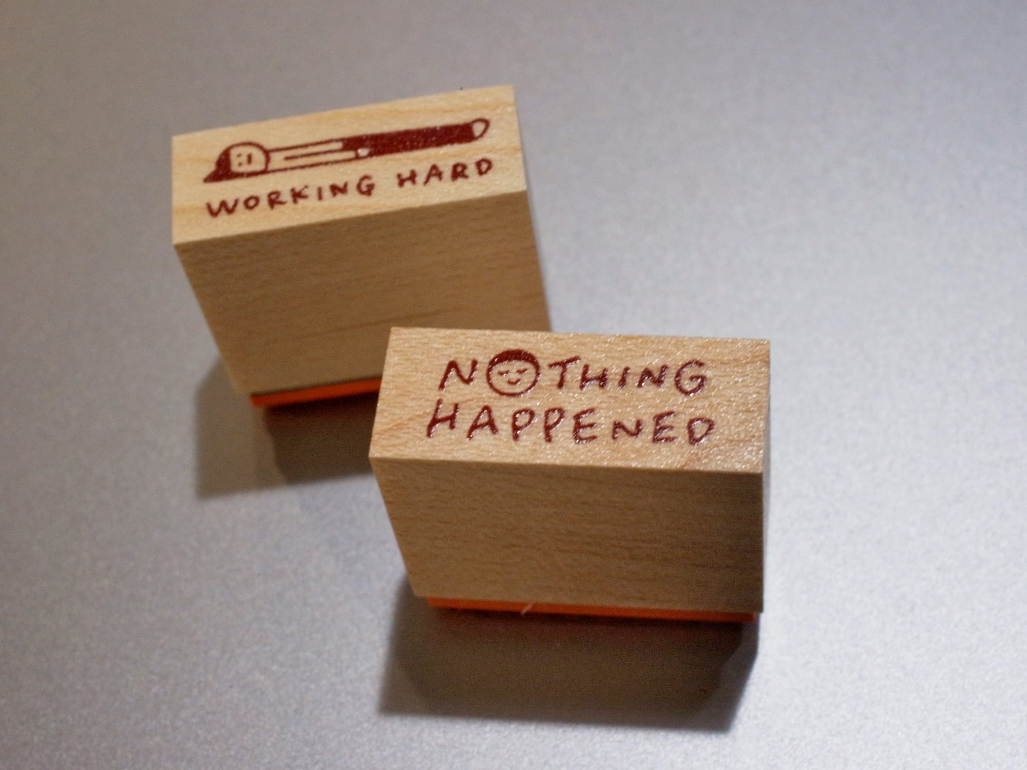 2 small rubber stamps with wooden handles: one is text, "nothing happened" with the "o" replaced by a smiling/smirking face, and the other is the same illustration of a person lying flat on the ground with the caption, "working hard"