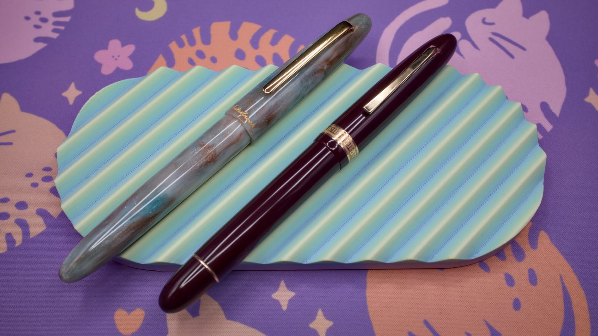 Two cigar-shaped fountain pens, one with a swirly, light-colored shimmery acrylic body, the other a dark burgundy resin body, sitting on a pastel green and yellow pen tray