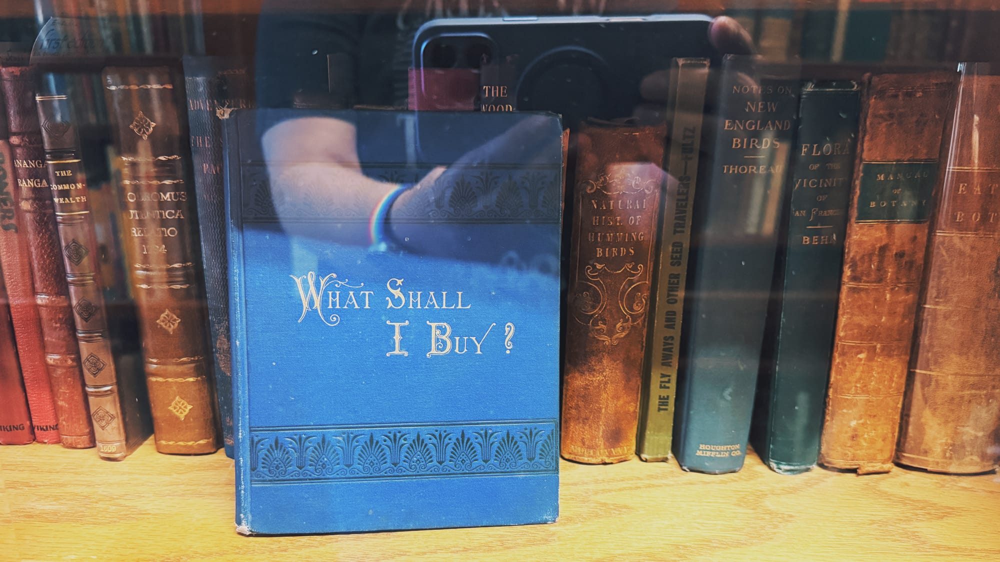 An old blue book titled, "What Shall I Buy?" sitting on a shelf behind glass in front of other old books shelved properly, a reflected image of hands holding an iPhone to take a picture overlaid, making the picture look like a double exposure