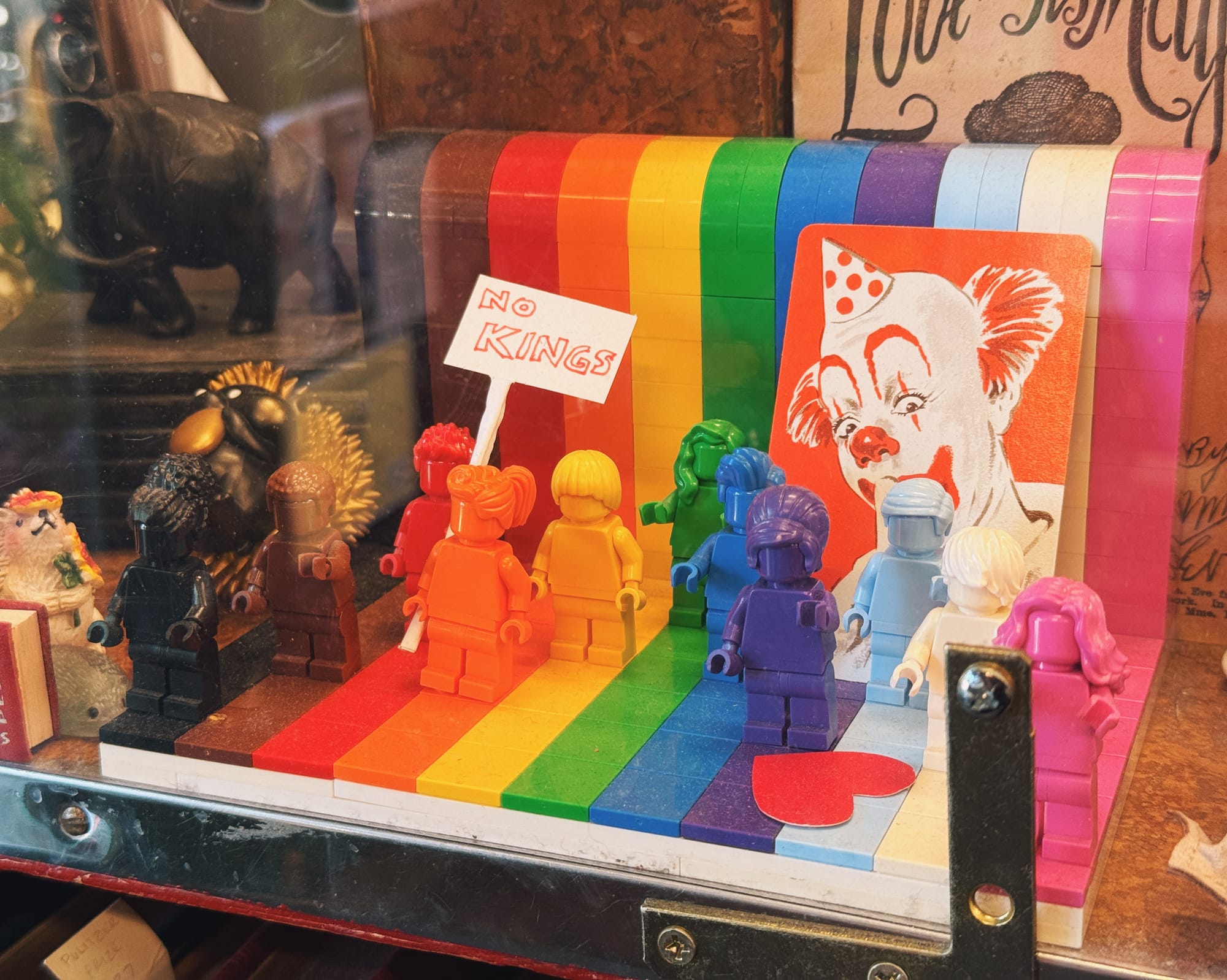 A small LEGO Pride set behind glass near the cash register showing minifigs in each color of the Pride flag, with an added "No Kings" sign placed between the red and orange minifigs, and randomly a picture of a clown propped up behind the blue, purple, and light blue minifigs