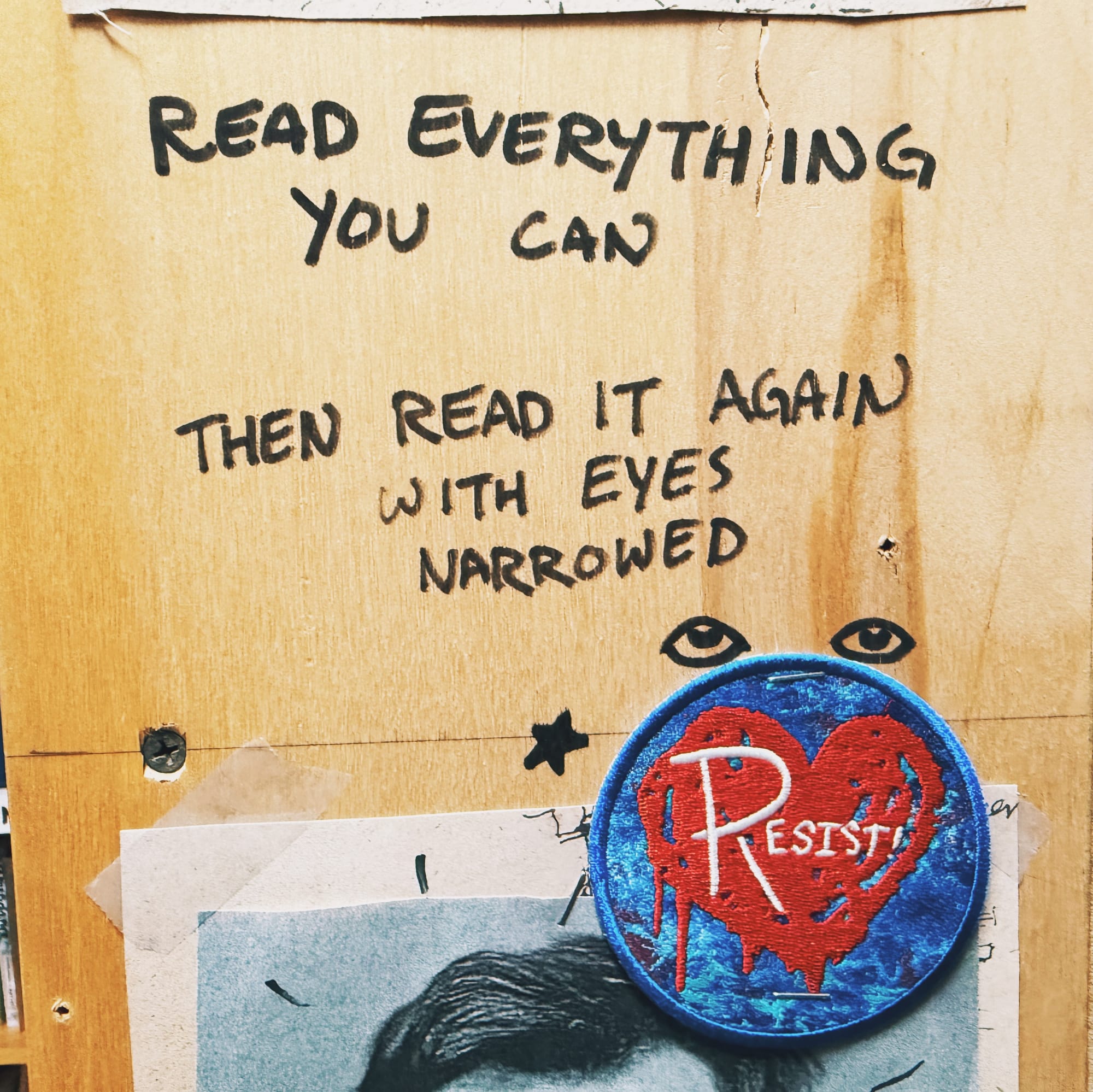 Text written on the side of a wooden bookshelf with black marker: "Read everything you can then read it again with eyes narrowed", and a blue circular patch with a red graffitied heart and "Resist!" in white on the heart stapled to the shelf with eyes and a star drawn above and to the left of the patch, respectively