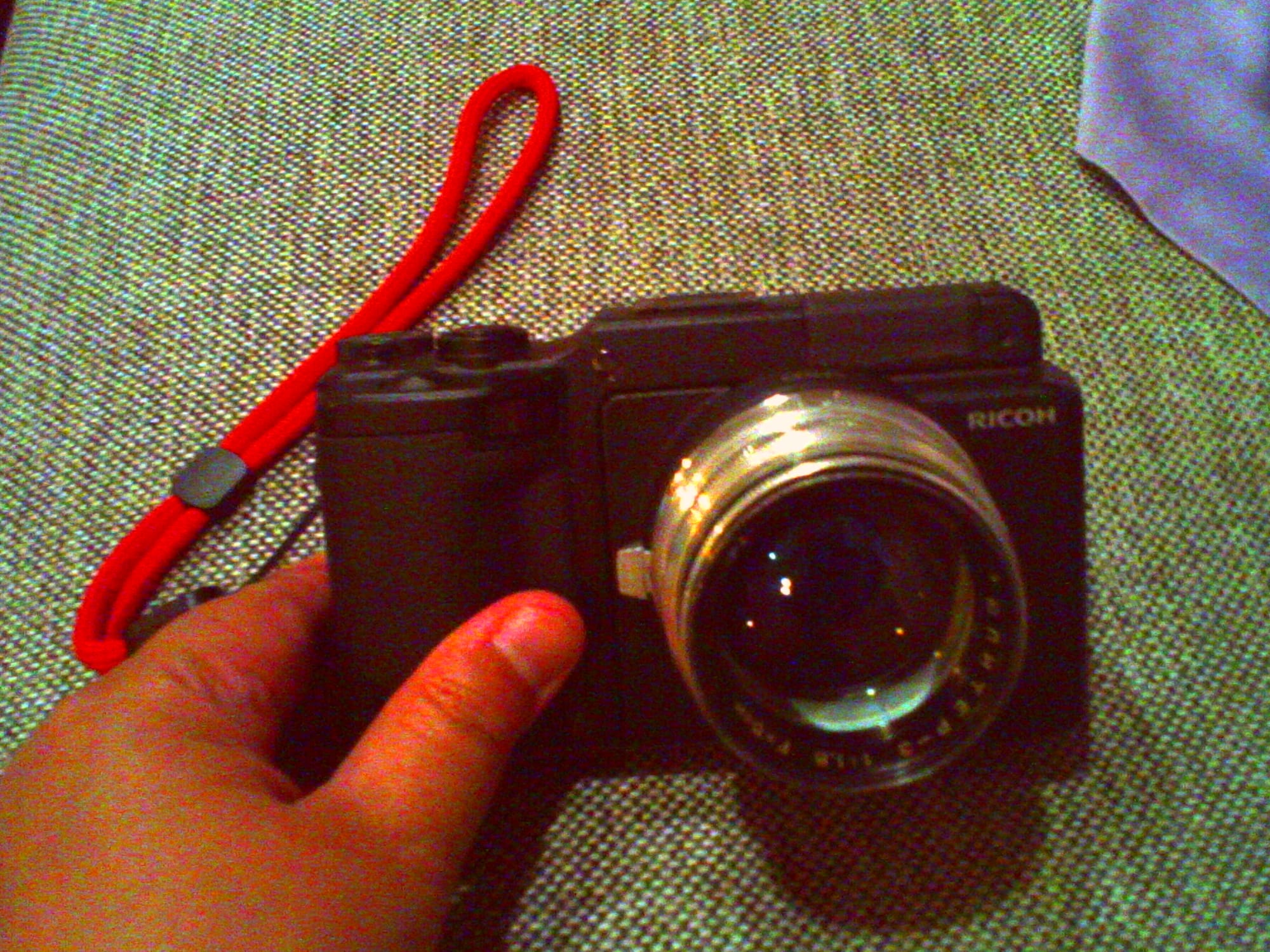 Very low-res, grainy, wildly-colored snapshot of a hand holding a Ricoh GXR up at an angle on a couch cushion