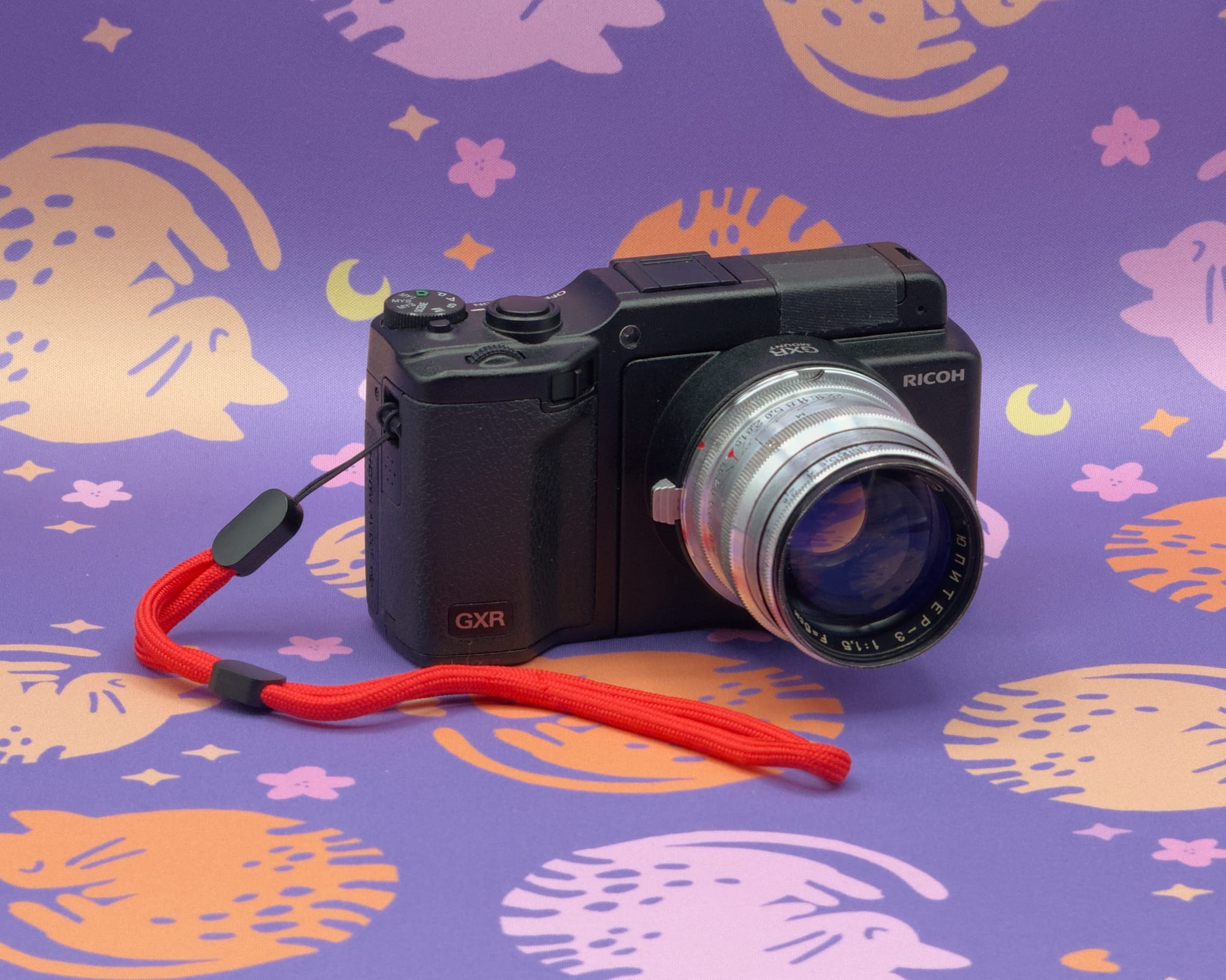 Black compact Ricoh GXR camera with a slightly scruffy, silver, vintage Russian lens attached, and a bright red wriststrap