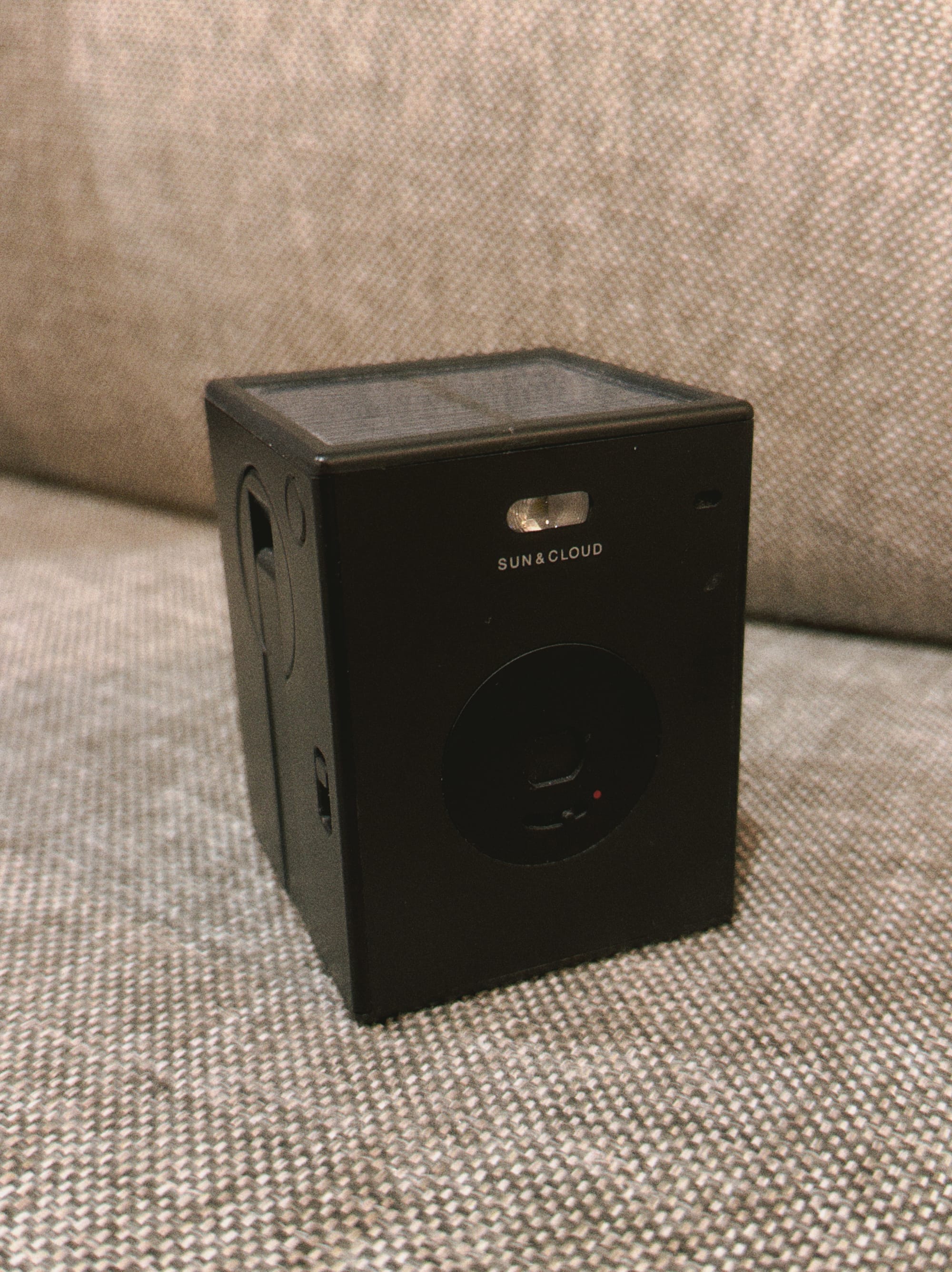 Black rectangular solid-shaped camera sitting on a gray sofa cushion, a solar panel visible on its top, and the outline of a folded away handcrank visible on its side