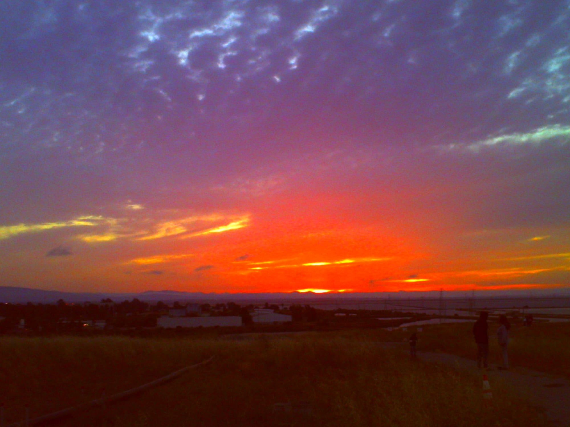 Overly saturated and low-res picture of the vivid red-orange sunset with a thick layer of clouds above it, and a dark horizon below