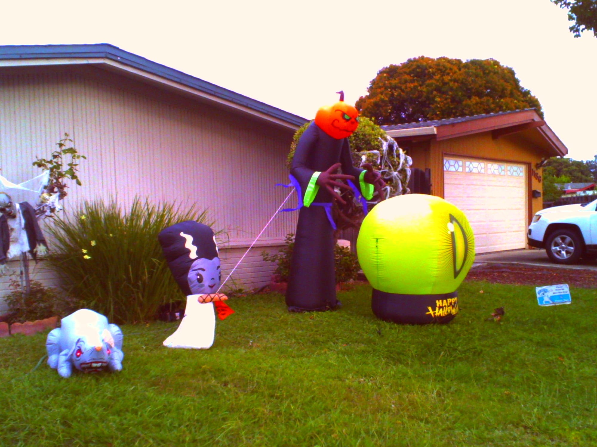 Low-res picture of a yard with various inflatable Halloween decorations, like a huge, red-eyed rat; a purple-faced person with a white dress and tall black hair with two white stripes; a tall person with an evil pumpkin head and black robe, and a large green crystal ball thing with an evil eye in the middle