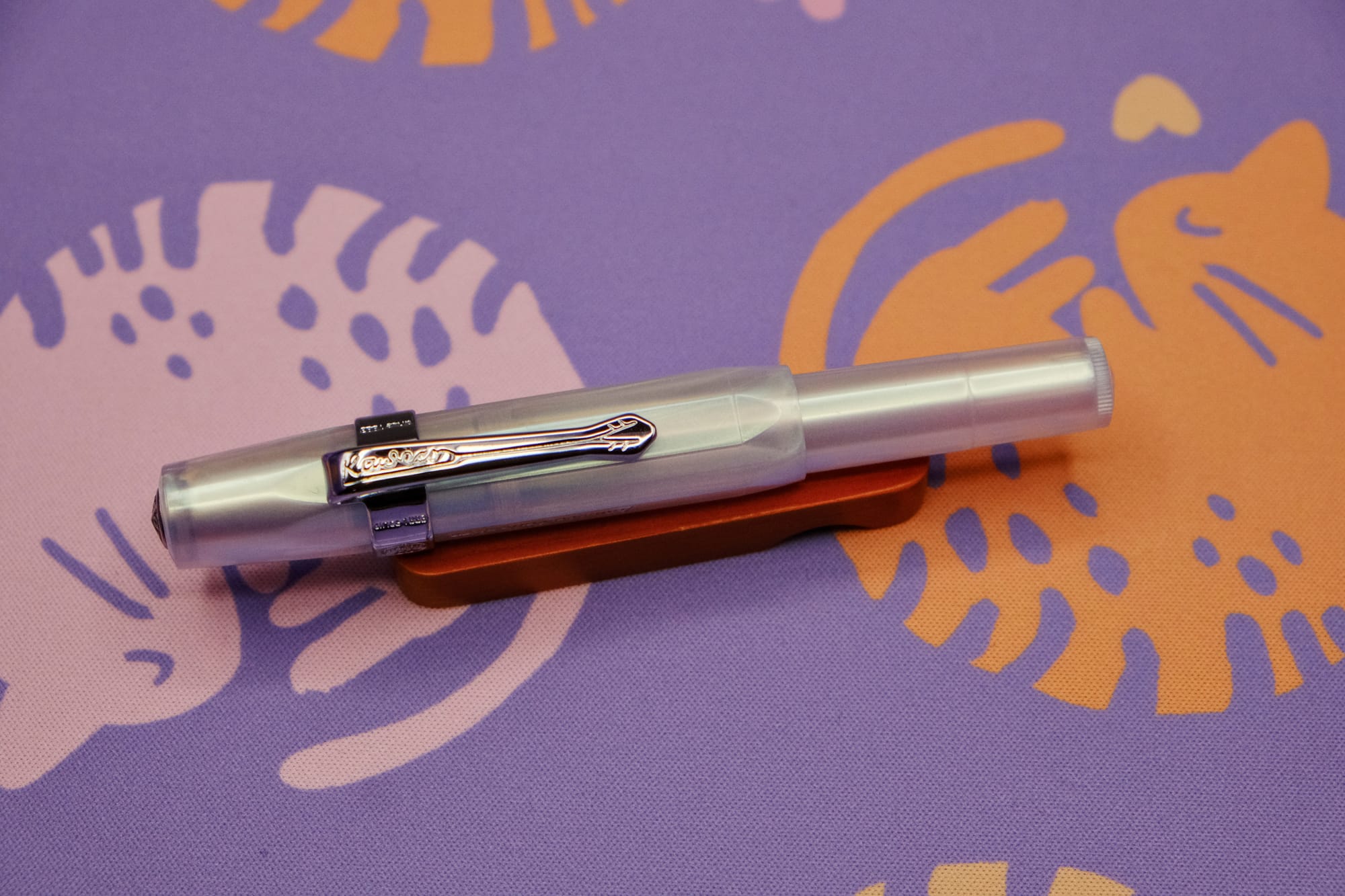 An iridescent pearl, plastic, pocket-sized fountain pen with a silver clip sitting on a wooden pen rest, with a purple deskmat with pink and orange curled up kitty pattern underneath